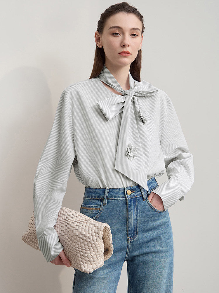 A woman modeling a light gray long-sleeve blouse with a large bow at the collar and decorative brooch details, paired with blue jeans and holding a beige woven clutch against a plain light gray studio background.