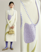 Woman wearing a long cream sweater with a purple floral pattern, holding a gold handbag.