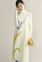 Woman wearing a long cream dress with green leaf patterns and a purple top, holding a gold handbag.