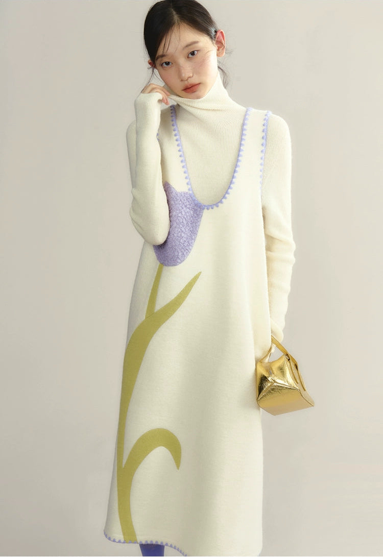 Woman wearing a long cream dress with green leaf patterns and a purple top, holding a gold handbag.