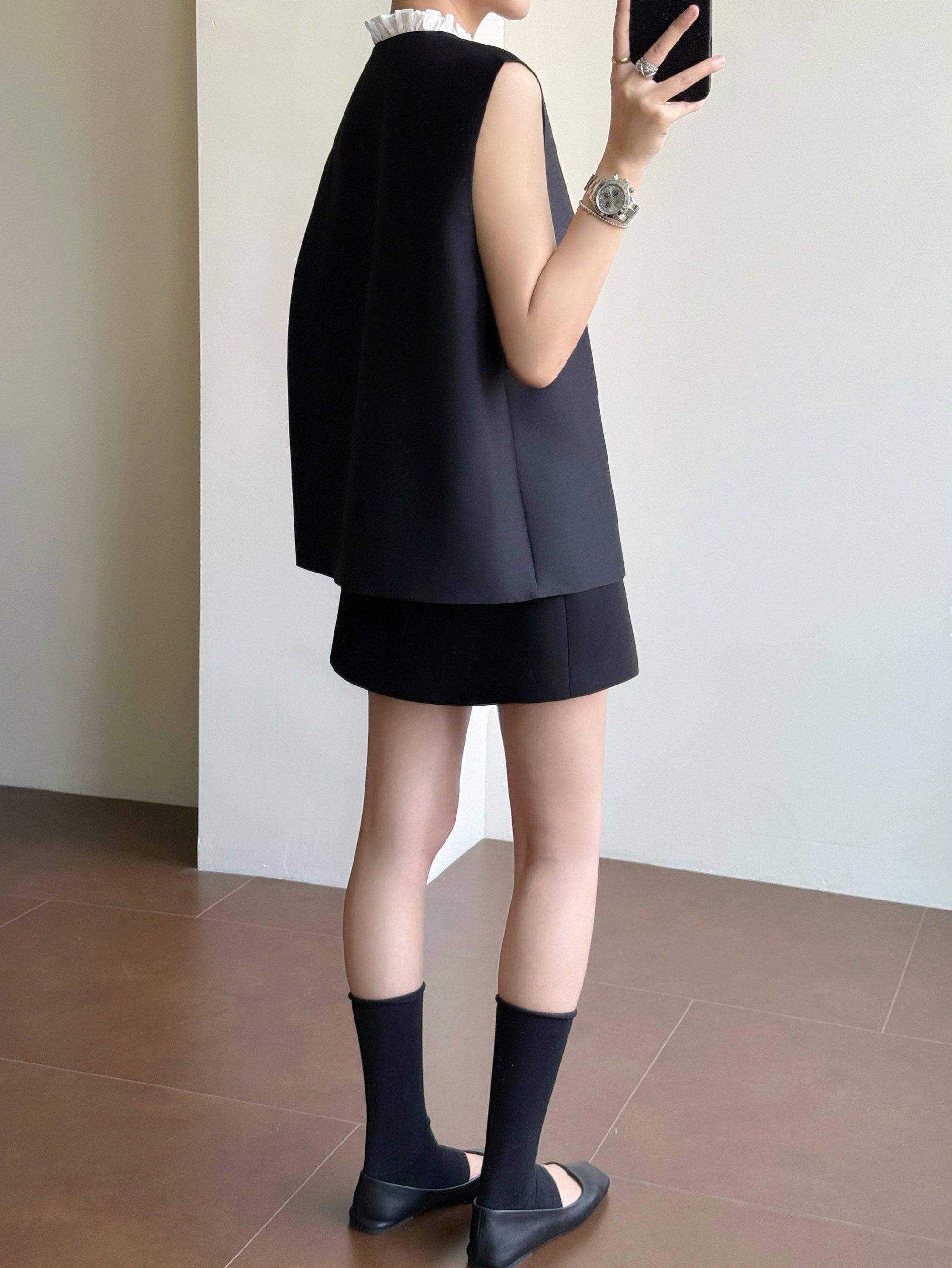 A model is shown from the back wearing a black sleeveless vest and matching black skirt, paired with black socks and black flats, standing in an indoor setting with a tiled floor and light-colored walls.