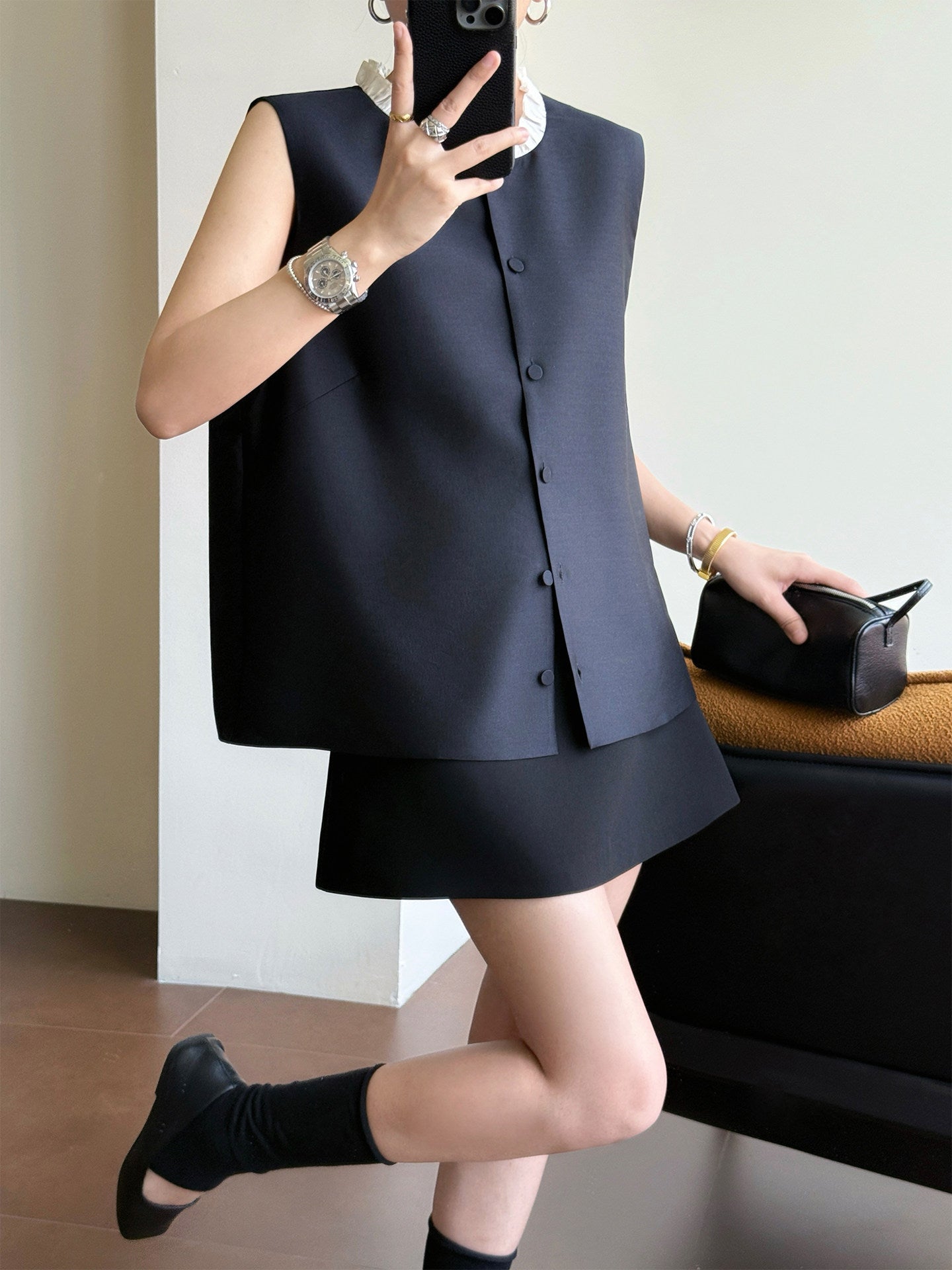 A person takes a mirror selfie wearing a dark navy sleeveless button-front vest and matching skirt set, with black socks and a black handbag nearby in an indoor room.