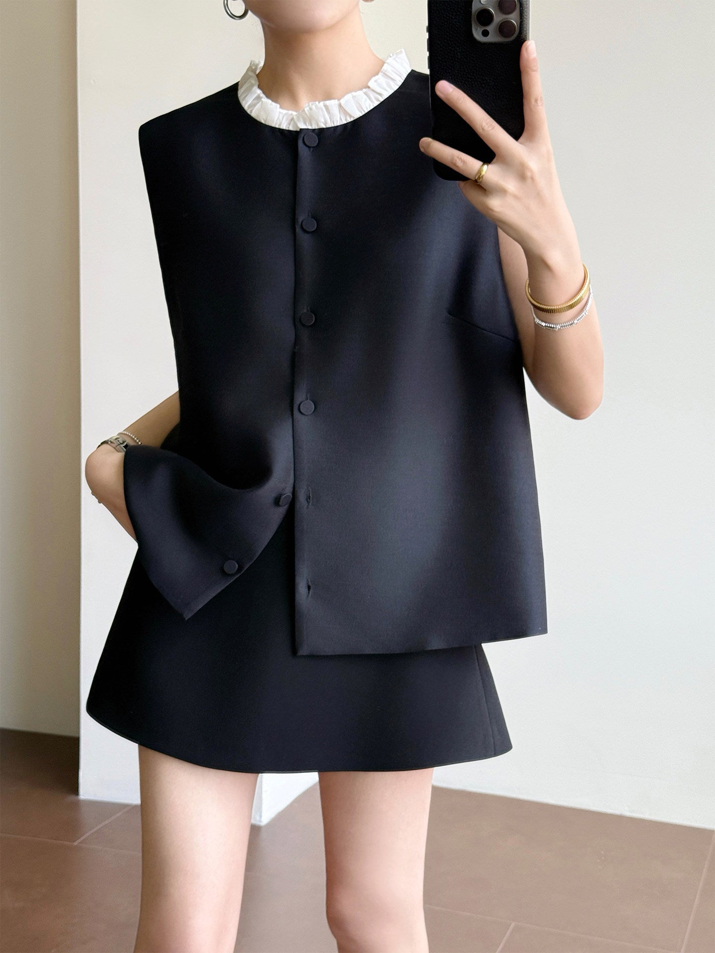 A woman takes a mirror selfie wearing a black sleeveless vest with a white ruffled collar and a matching black skirt. She is holding a black smartphone to take the photo in an indoor setting with a light-colored wall and tiled floor.