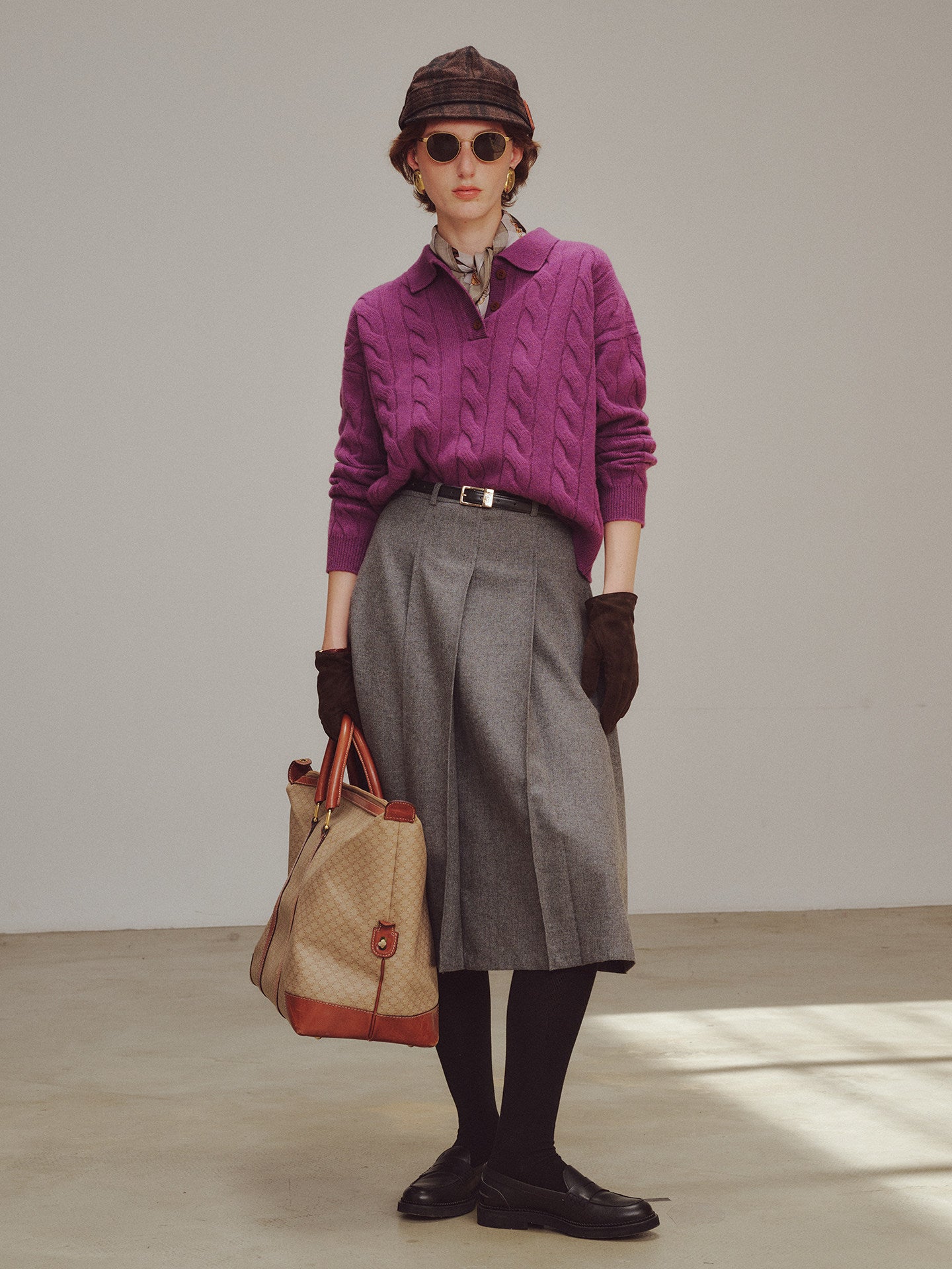 Person wearing a purple sweater, gray skirt, and holding a beige handbag against a neutral background