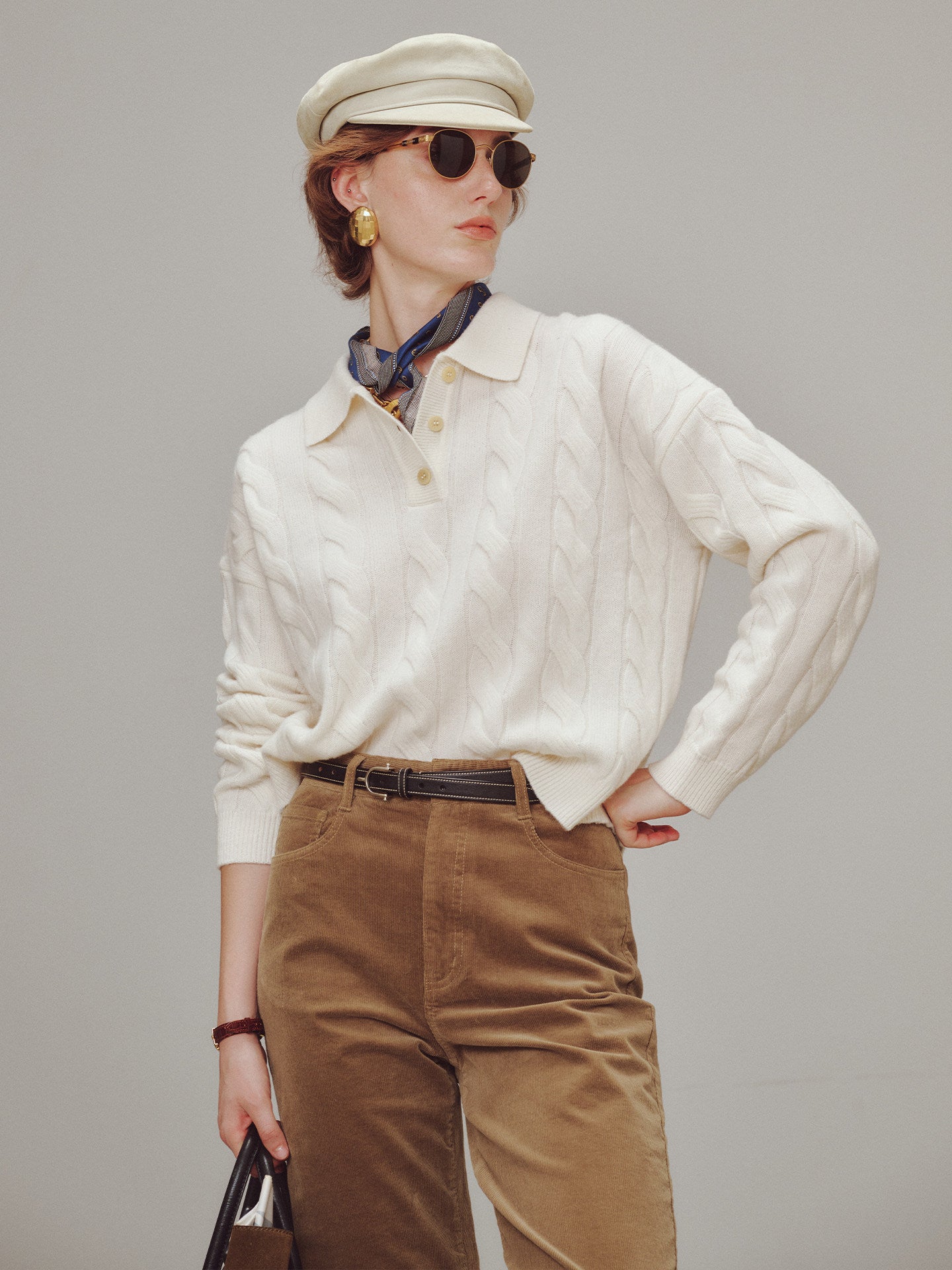 Woman wearing a cream sweater, beige cap, sunglasses, and brown pants against a gray background