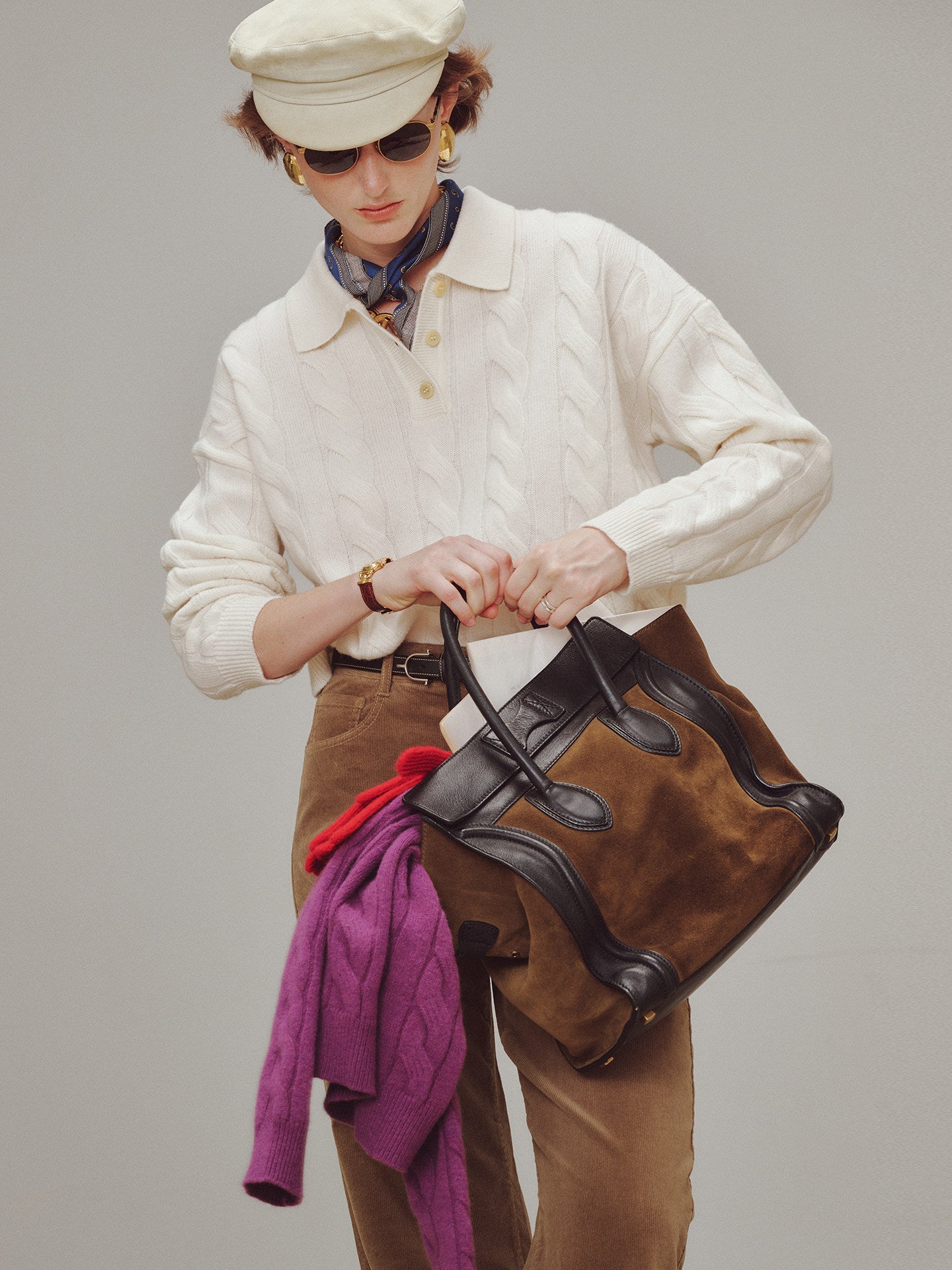 Person holding a brown leather bag with a purple sweater over their shoulder against a gray background