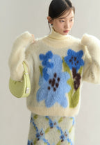 Woman wearing a cream sweater with floral patterns, holding a green handbag against a plain background