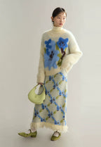 Woman wearing a cream sweater with blue floral patterns and a matching skirt, holding a green handbag.