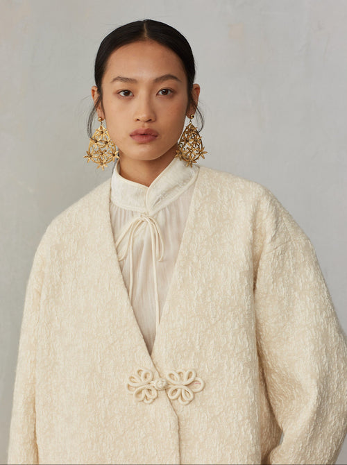Woman wearing a cream-colored textured coat with floral details on a plain background