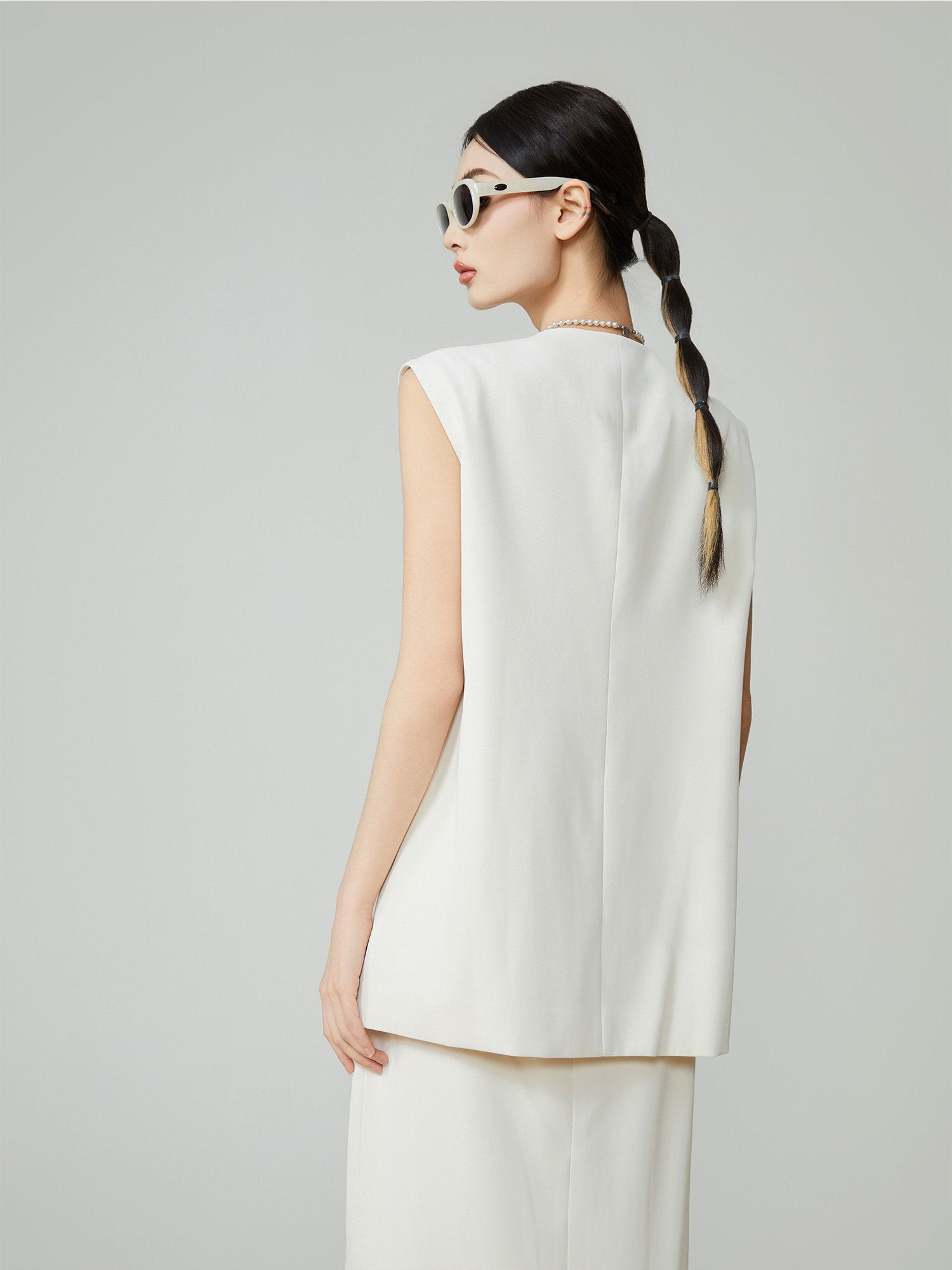 Woman wearing a white sleeveless top and pants with sunglasses on a gray background