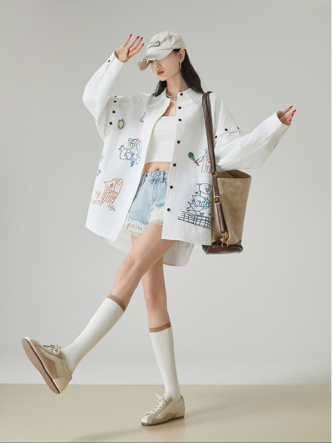 Woman wearing a white embroidered jacket and denim shorts with a beige bag, standing against a plain background.
