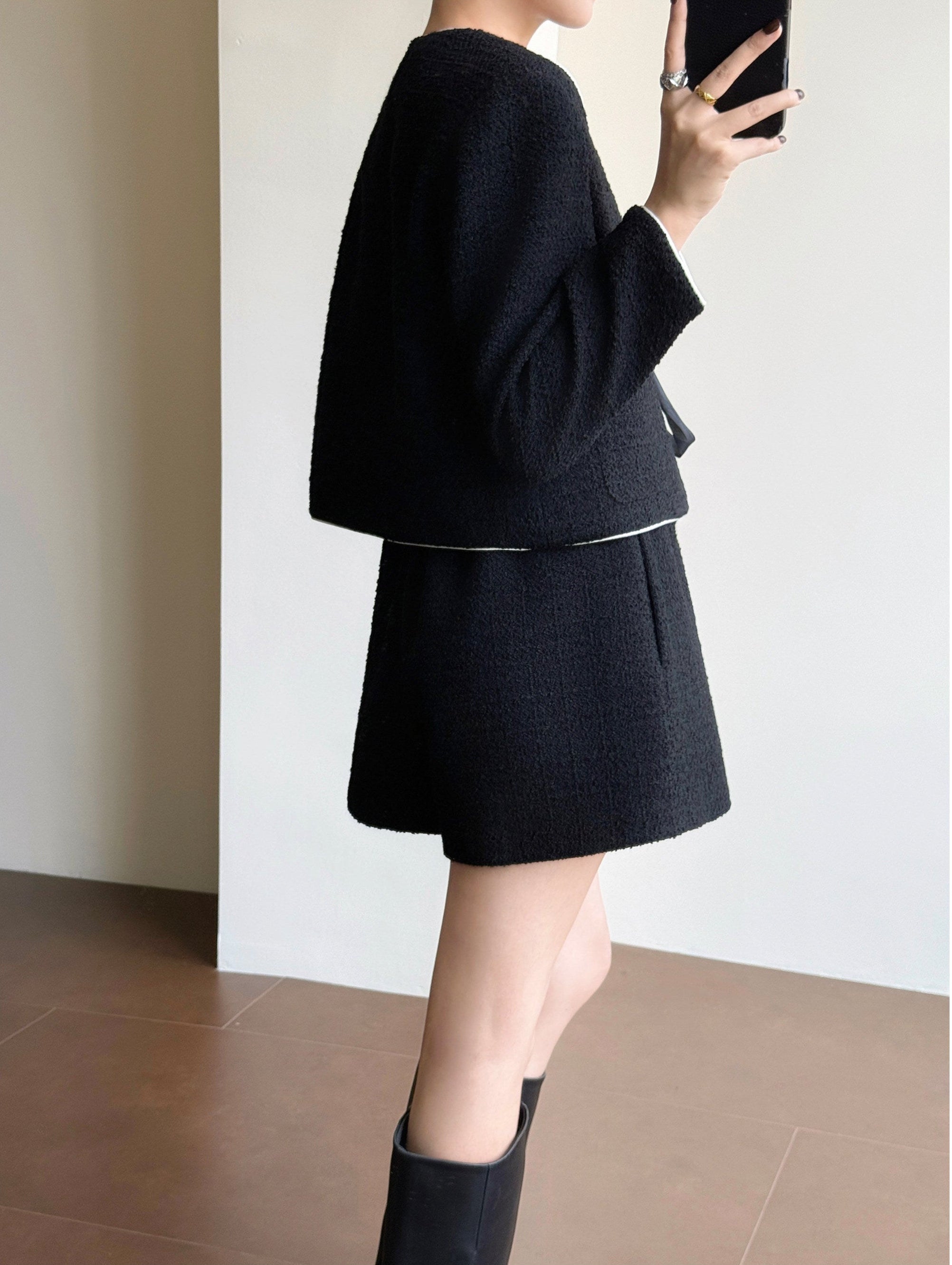 A person shown from the side in a black textured knit two-piece outfit consisting of a cropped long-sleeve top and a matching short skirt, paired with black ankle boots, standing indoors against a light wall.
