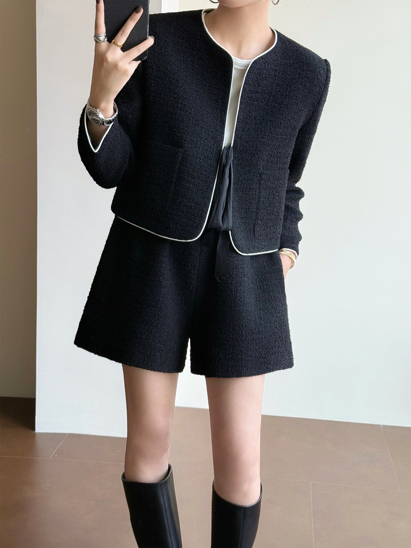 A person wearing a black tweed short jacket and matching shorts with thin light piping trim, paired with a white top and black knee-high boots, photographed indoors against a plain light wall.