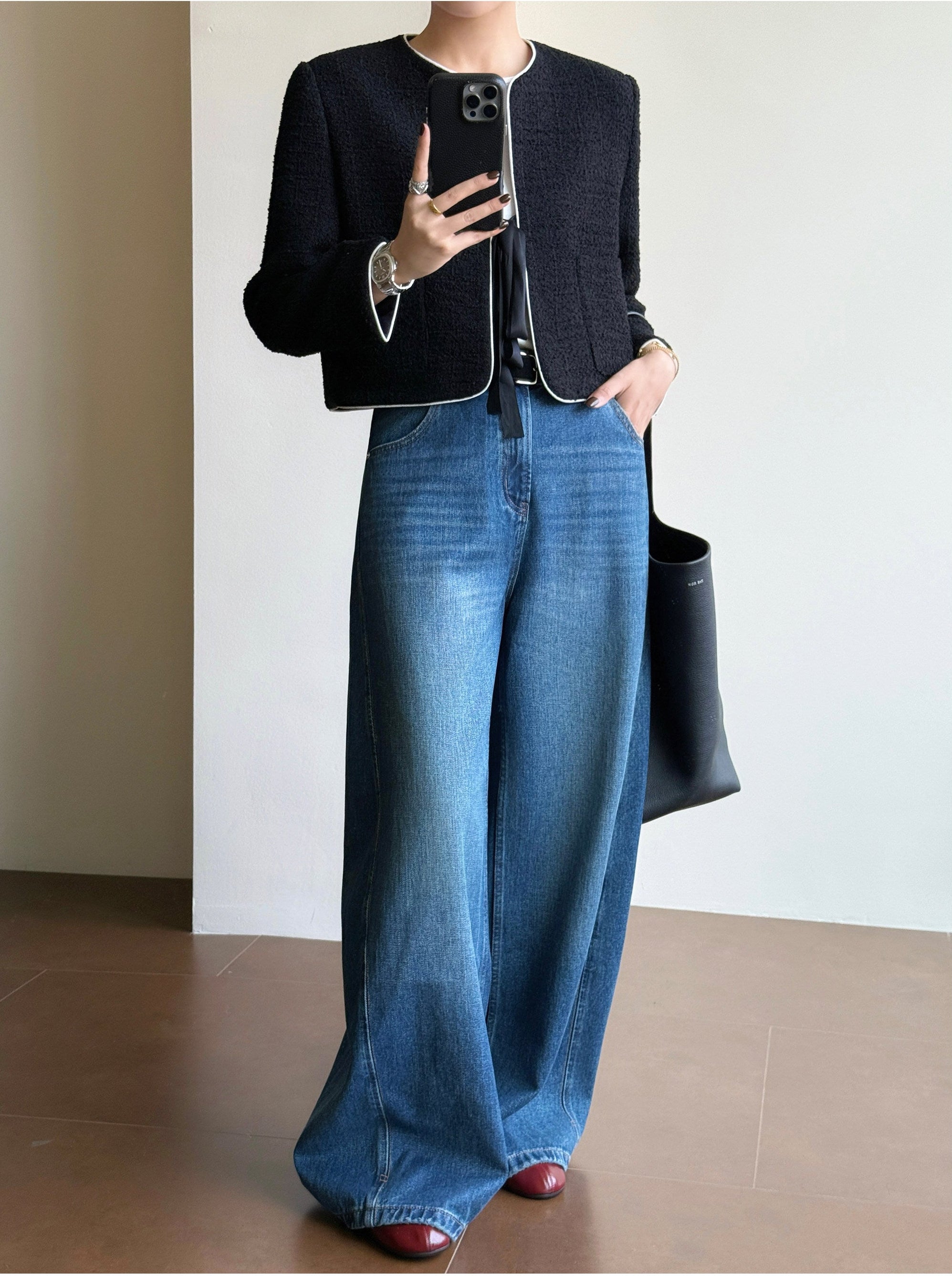 A person modeling a black textured cropped jacket with white piping and wide-leg blue denim pants, holding a black tote bag in a minimalist indoor setting.